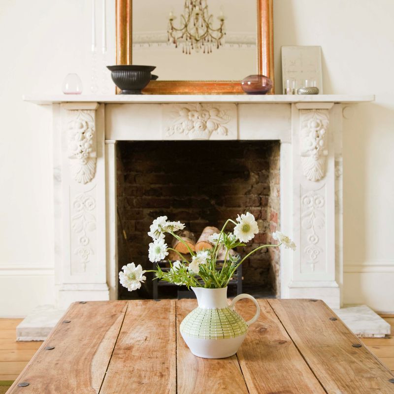 a white old-fashioned-looking fireplace with a wooden table with a vase of flowers in front of it